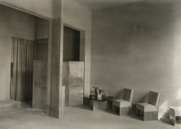 Boutique interior late 1920s. Black- and white photo. Chairs against a wall and a folding screen in aluminium. Flowers on a coffee table.