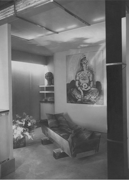 Bedroom 1920s style, black and white photo. A bed made of glass that resembles a coffin with out lid in the middle of the floor. A big oil painting on the wall and a bust of a woman on a shelf.