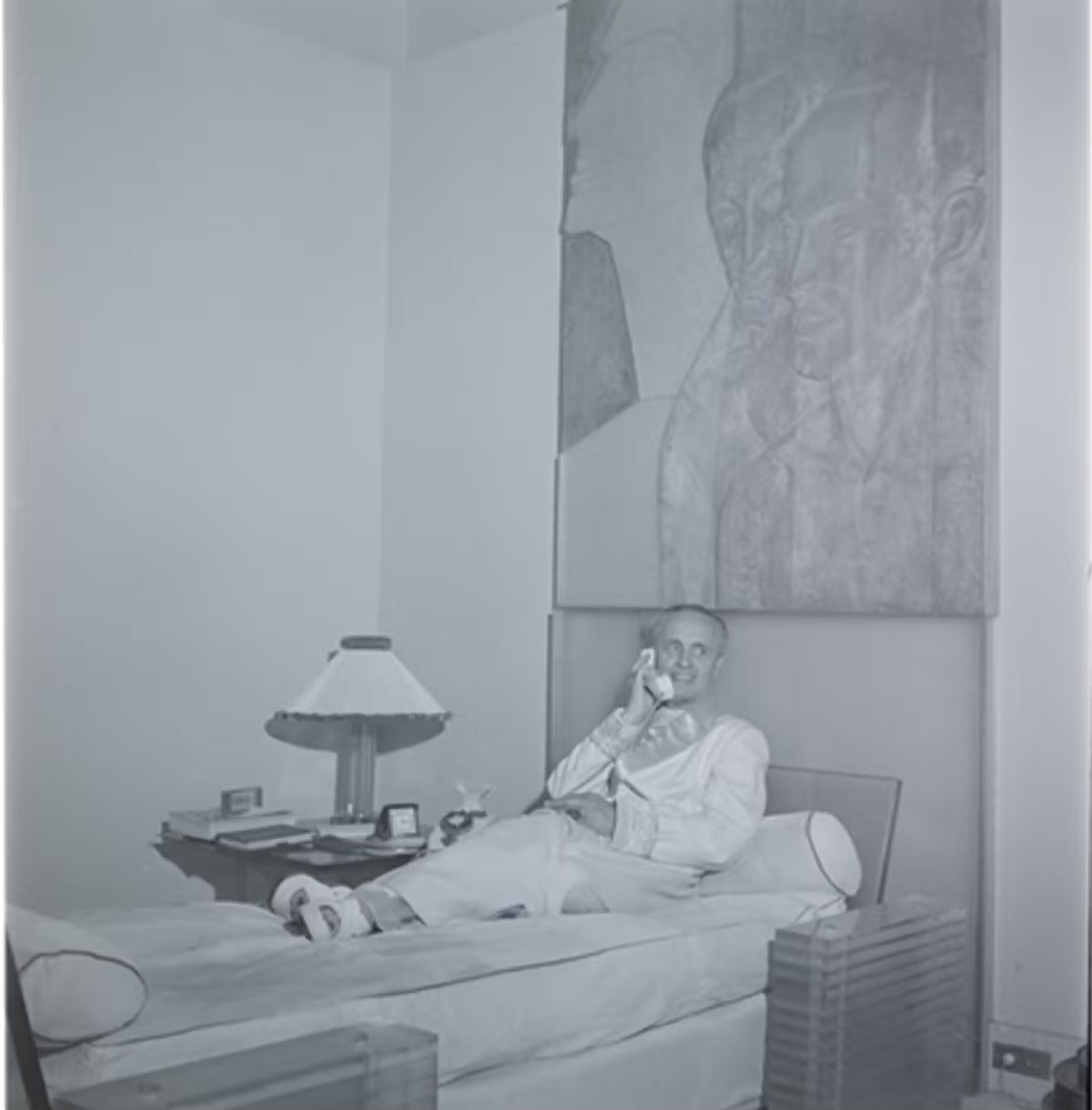 Man in bed talking on the phone, 1920s style, black and white photo. Big oil painting on the wall behind him,