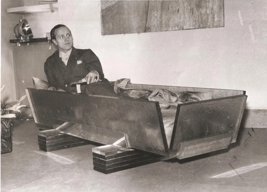 Bedroom 1920s style, black and white photo. A man in a bed made of glass that resembles a coffin with out lid 