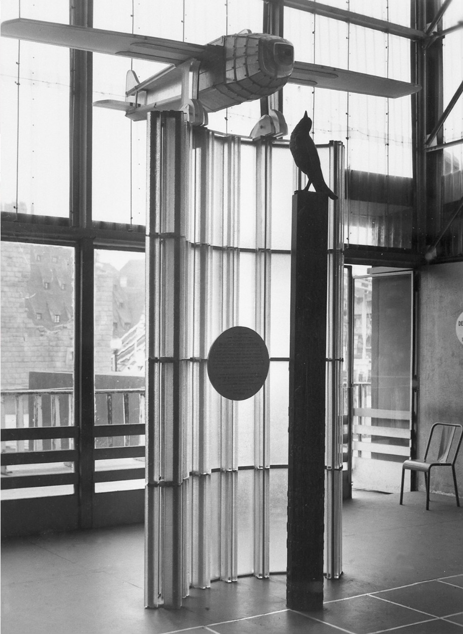 Modernist exhibition interior featuring a glass and aluminium folding screen, a suspended airplane sculpture, and a bird sculpture on a pedestal, emphasizing light, transparency, and geometric form.